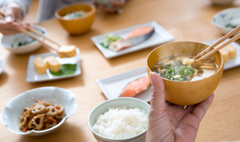 A hand holding a bowl of miso soup and a table with many japanese dishes laid out